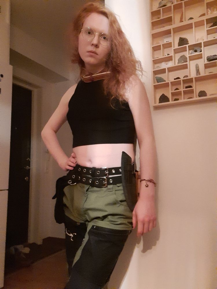 Me standing against a corner with an arm on my hip. I'm dressed in a black croptop, green and black pants, a collar, utility belt with two belts, which has two knives (one of which is a set of three) attached to it. I'm looking down a little at the camera