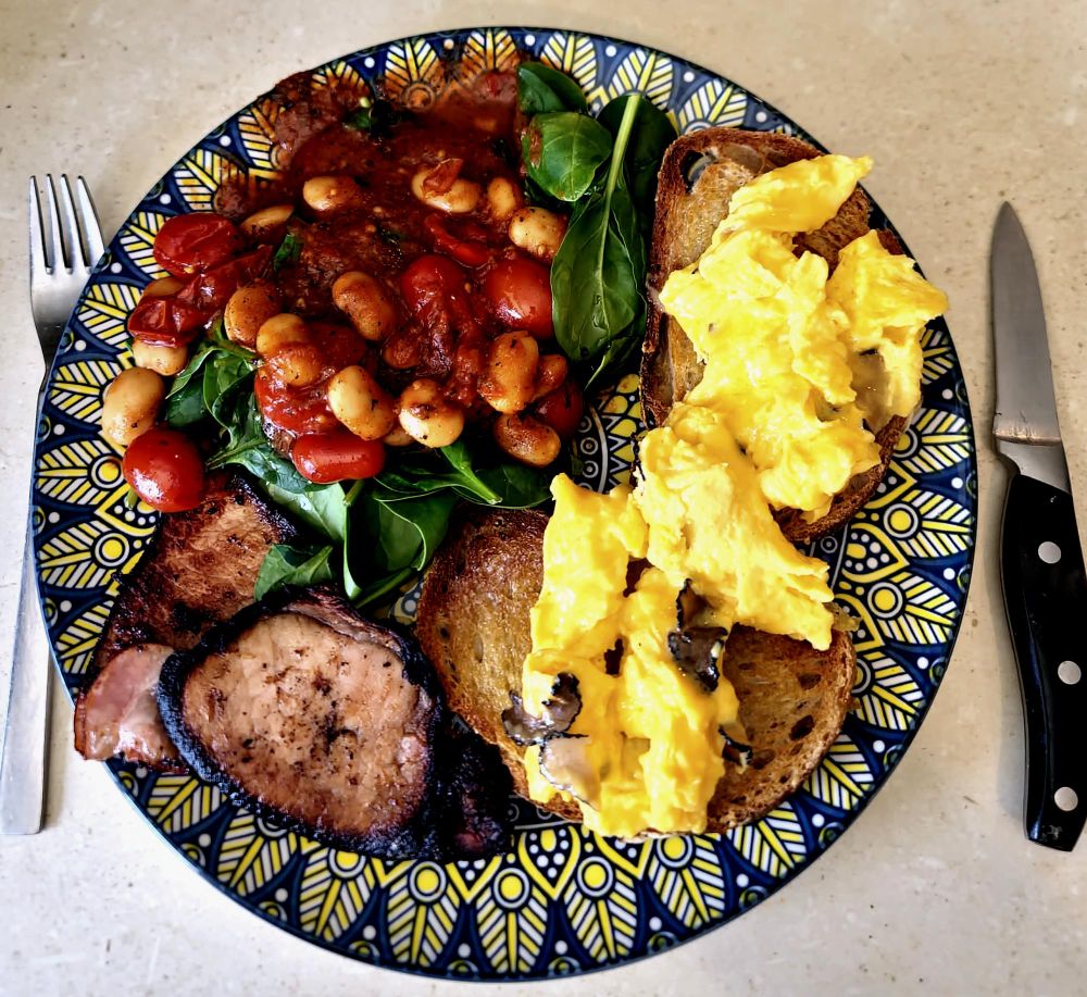 On the photographer's kitchen bench is a blue and yellow plate. On it are two pieces of light rye sourdough toast topped with scrambled eggs with tiny truffle slices. There is also some bacon, and a salad of fresh baby spinach leaves and cooked cherry tomatoes and cannelini beans.
