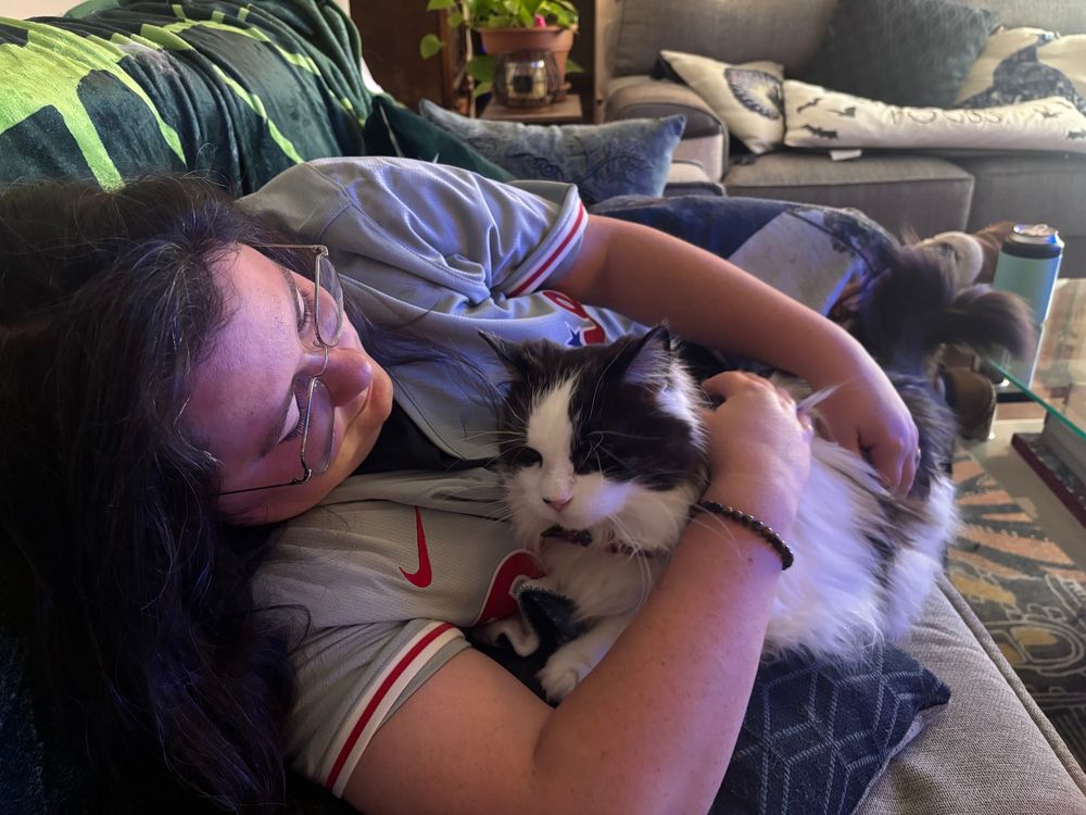 A woman laying down on the couch in a gray Phillies jersey. She is hugging a small black and white longhair cat.