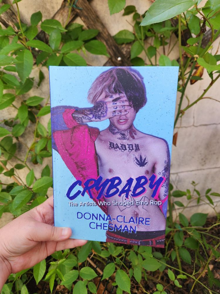 A hand holding a blue and pink book in front of a rose bush. The book is CRYBABY: The Artists Who Shaped Emo Rap.
