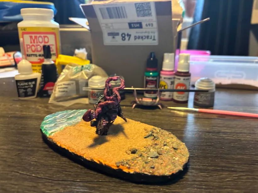A zoomed out photograph of a painted modded Warhammer 40k model on a custom made base made to look like a beach, in the background are glues, basing materials, paints, and a handy lil crab holding up a paintbrush, as well as a tub of sand and other miscellainy.