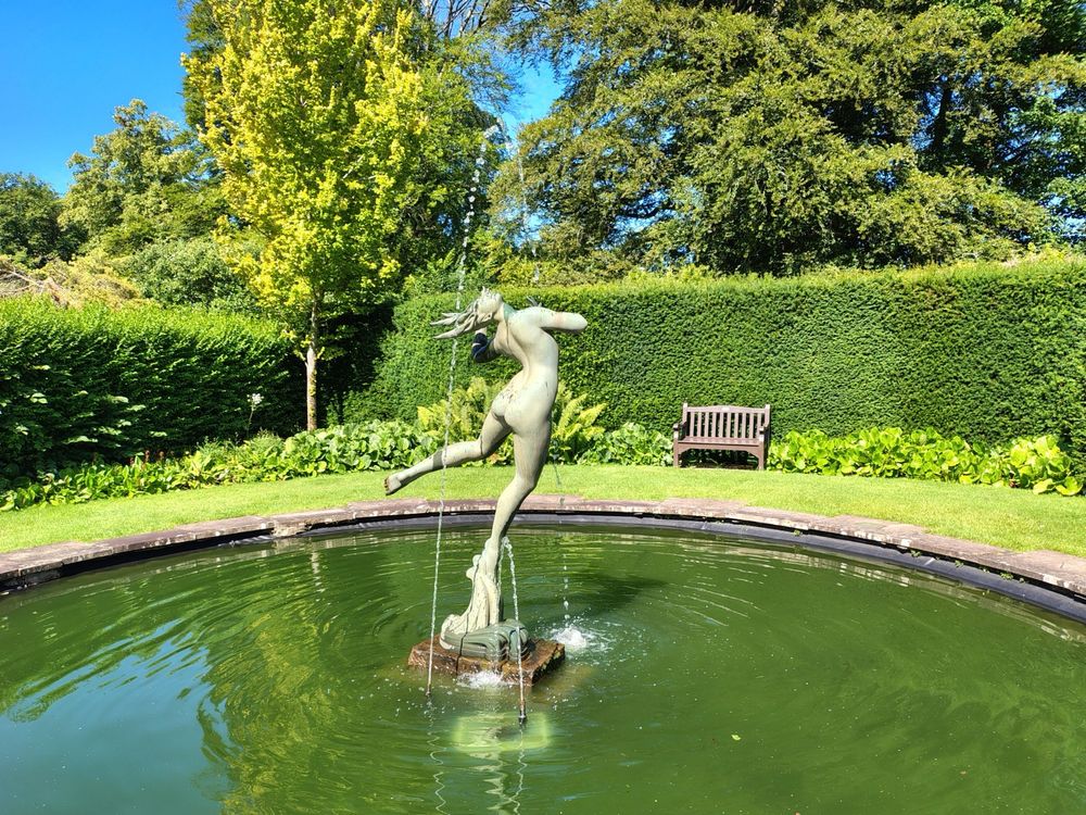 Another picture of the statue, Foam, at Greenbank Gardens, standing in the centre of a pool of water. Blues skies and vegetation visible in the background.