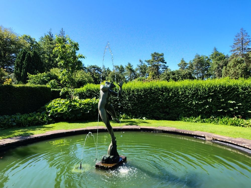 Picture of the statue, Foam, at Greenbank Gardens, standing in the centre of a pool of water. Blues skies and vegetation visible in the background.