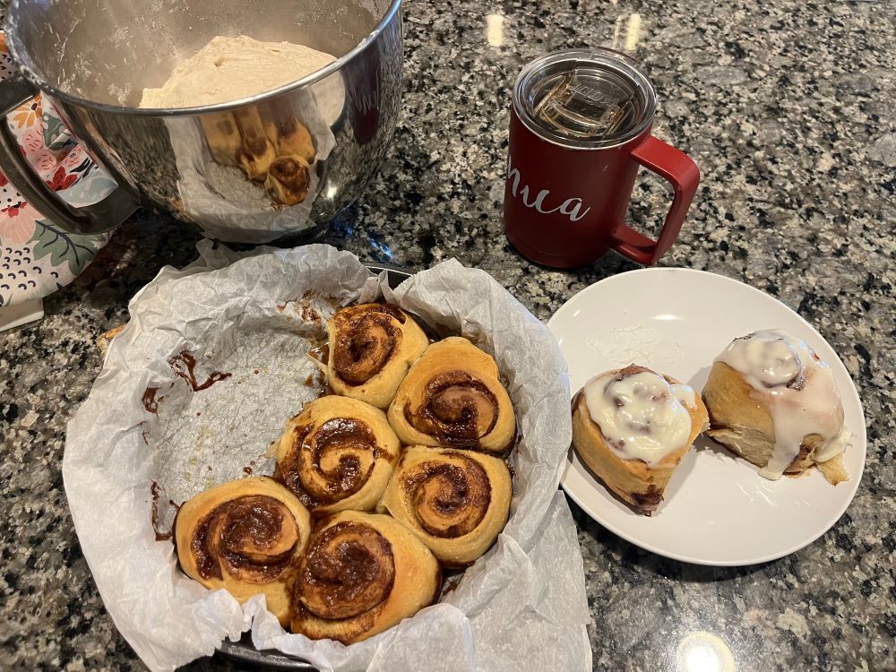 Left to right: top: stainless steel bowl with sourdough proofing, red cup of coffee. Bottom: pan of fresh baked cinnamon rolls, plate with two cinnamons with icing drizzled on top.