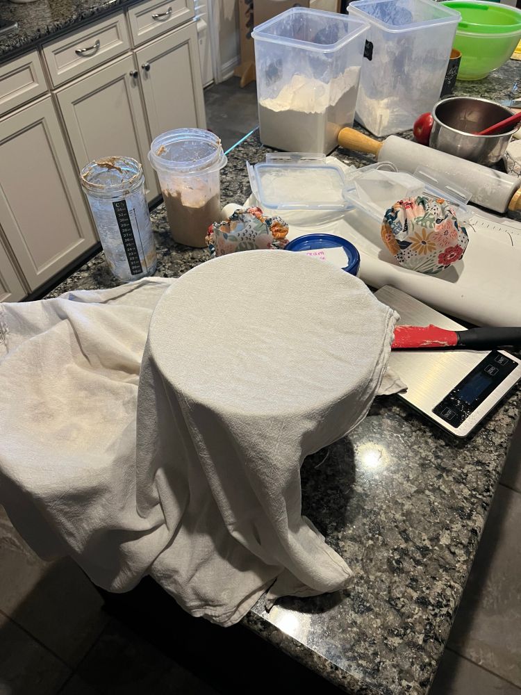 Granite countertop with baking mess: bowls covered in towels, scale, dirty spatula, open sourdough starters, 2 open containers of flour, folded over rolling mat, marble rolling pin, more bowls