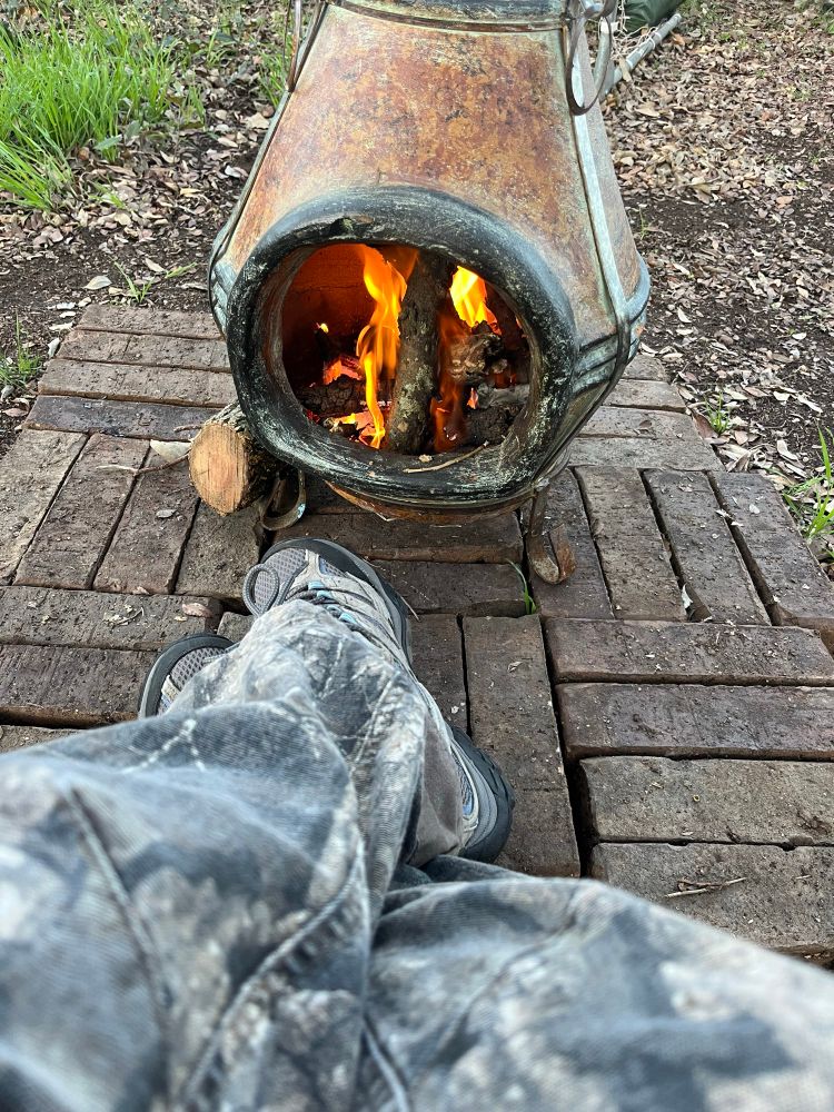 Two legs in camo pants extend to the foot of a mini chiminea with a glowing fire inside. Morning light.
