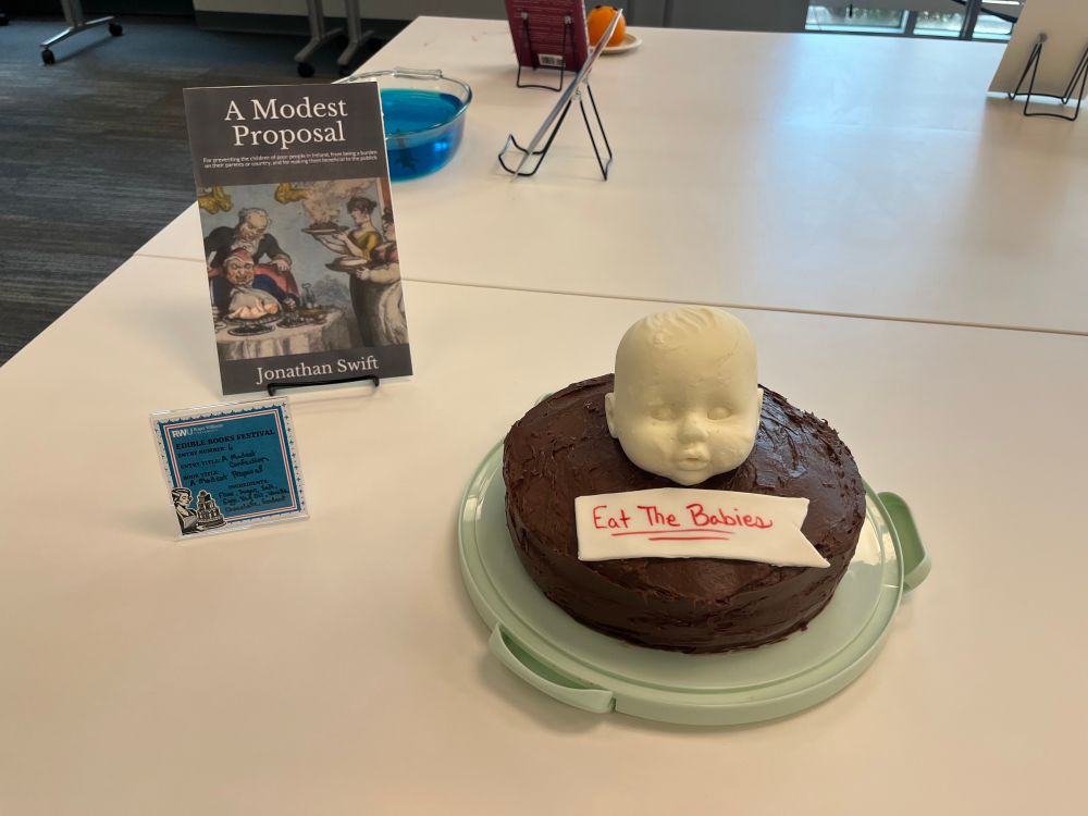 A double layer yellow cake with chocolate frosting and a solid white chocolate baby head with a fondant sign that reads Eat the Babies! In red edible ink as a nod to Swift’s A Modest Proposal.