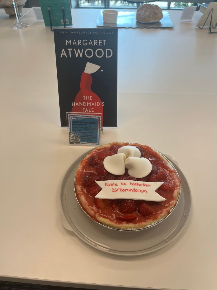 A Strawberry pie topped with three fondant hats and a sign that reads Nolite te Bastardes Carborundorum in red edible ink, as a nod to the Handmaid’s Tale.