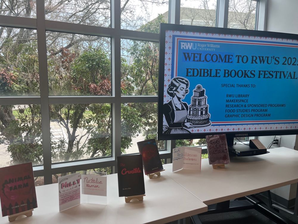 A digital screen reads “Welcome to RWU’s 2025 Edible Books Festival” with a vintage-style graphic of a woman holding a cake. Below the screen is a row of printed student-designed book covers inspired by food, including Animal Farm, The Crucible, and The Silence of the Lambs.