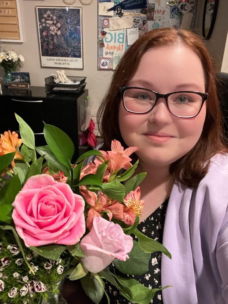 A librarian with dark hair, glasses, and a lilac blazer posing with a vase of cut flowers including roses and Calla lillies.