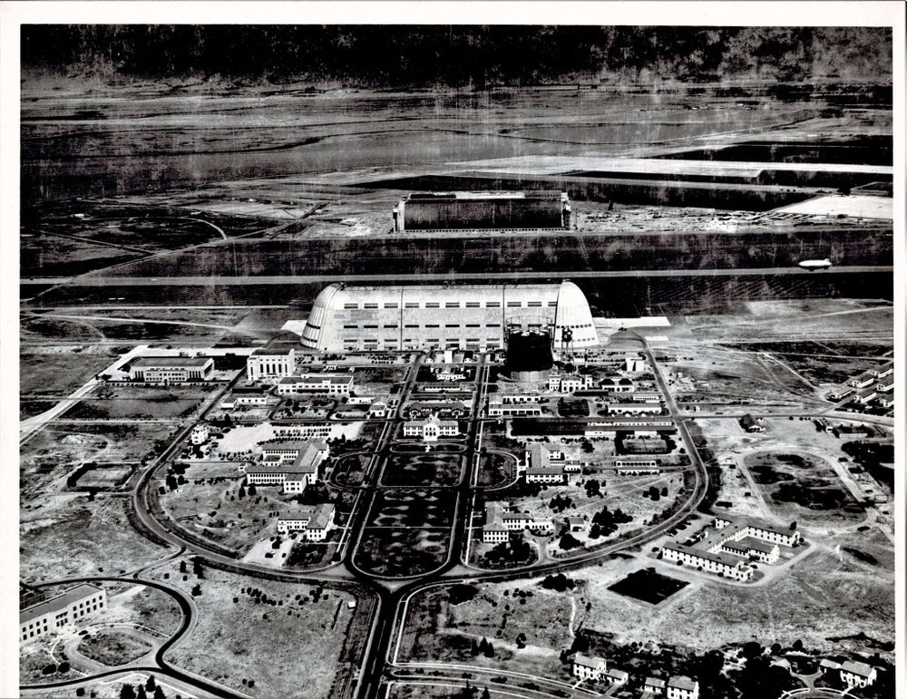 B&W aerial photo of KNUQ airfield, with Shenandoah plaza, hangar 1, the airstrip, and hangars 2 and 3 in the background.