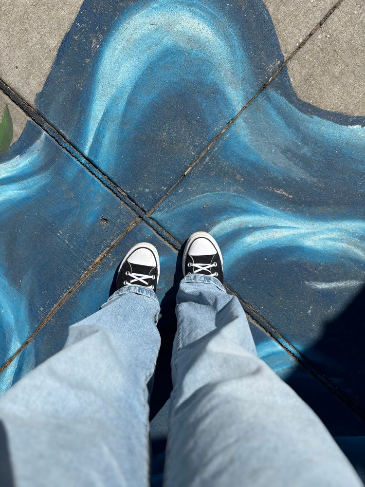 Photo looking down at a sidewalk painted to look like waves. A person wearing black and white Converse and wide-leg, light-wash jeans stands on the painted waves.
