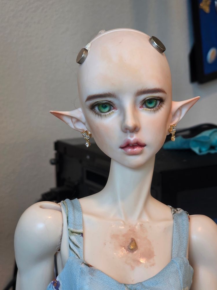 A doll shown from bust up without a wig. She has elf ears and there are magnets on her head