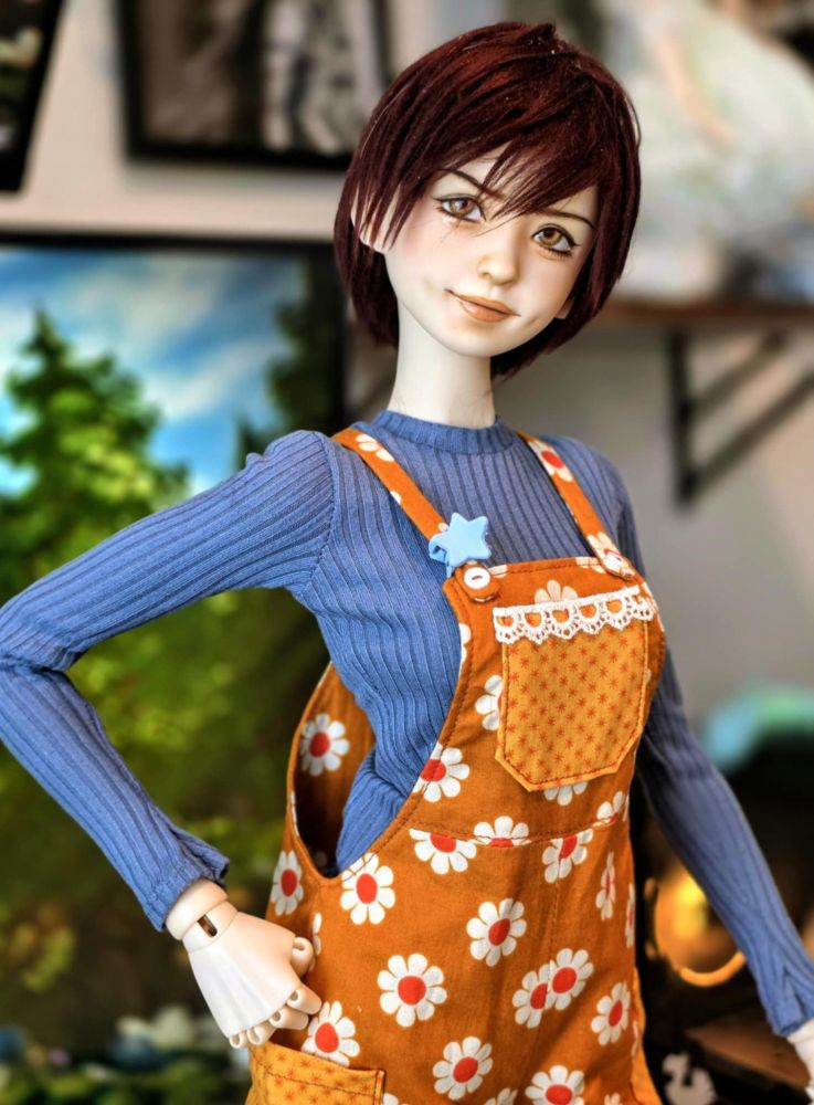 a doll posing on a desk in orange overalls and a blue shirt