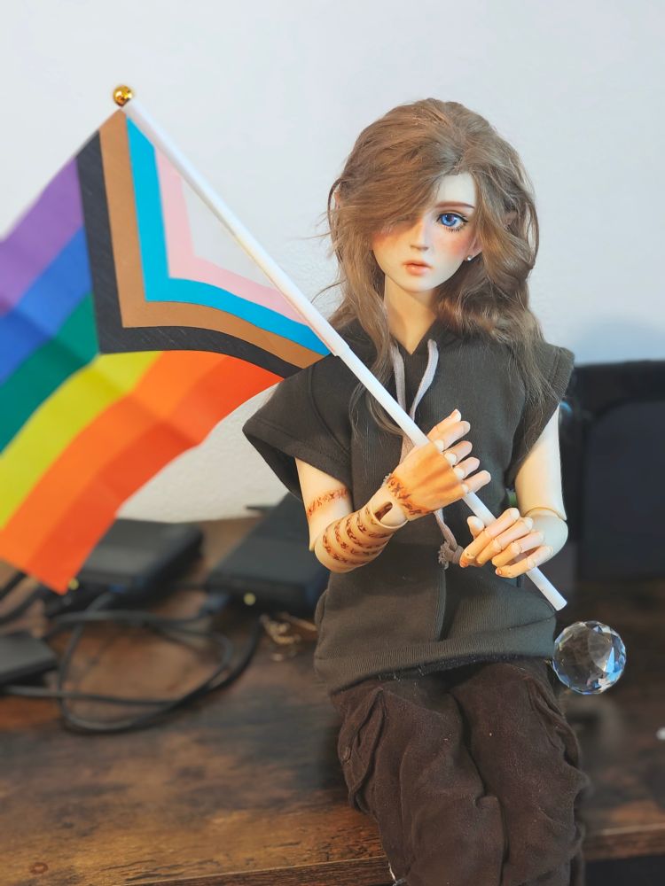 A doll with arm tattoos and fluffy long hair holding a progress pride flag, sitting on a table. They are wearing dark green and brown and look very androgynous 