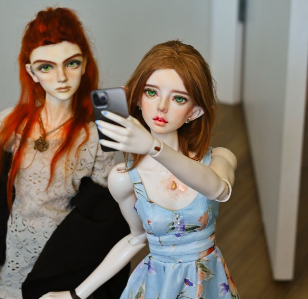 Two dolls posed taking a selfie with a miniature phone, shown from hips up. The one in front has elf ears and is wearing a blue dress. The one behind her has long red hair and is wearing a beige tunic top with a brown cardigan 