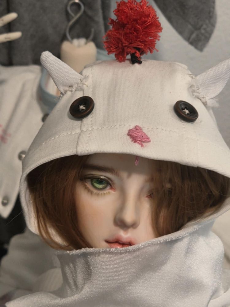 a close up of a doll's face. She is wearing a white hoodie resembling a Final Fantasy moogle, with the details on the raised hood. She is also wearing a scarf