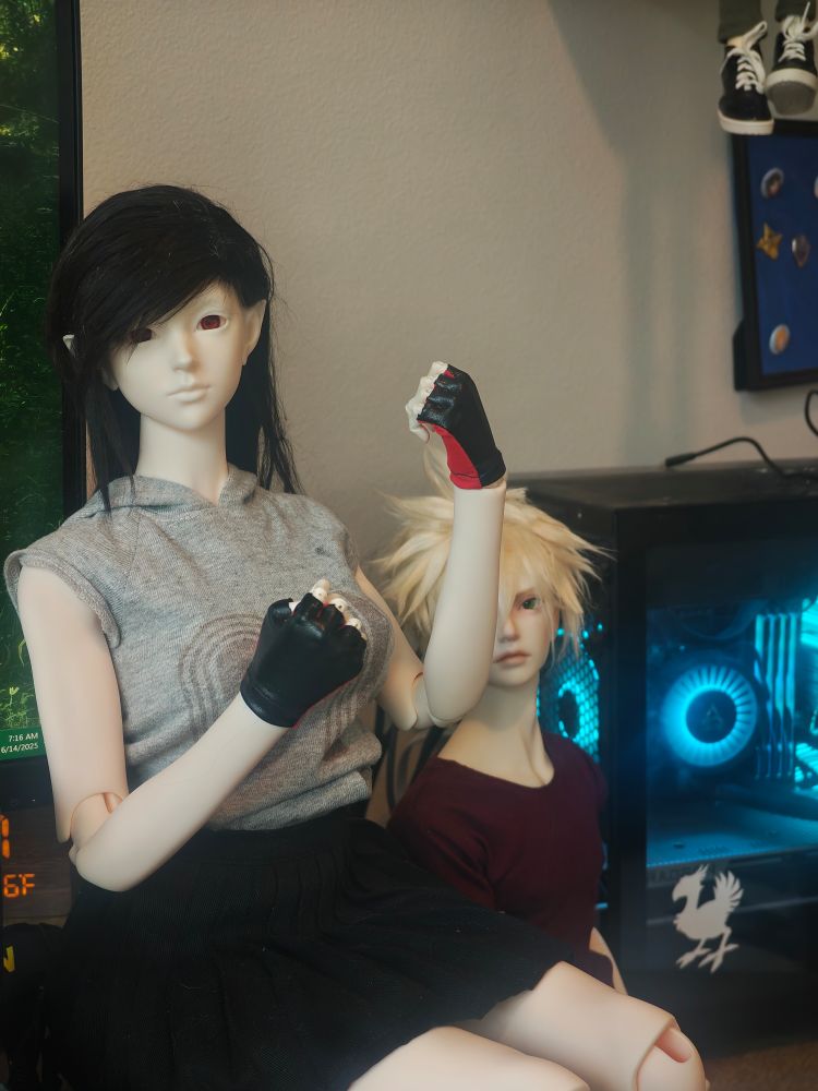 A doll wearing a grey shirt with her hands raised in a combative fist pose. She has black hair and is wearing fingerless gloves

Another blond doll is in the background, along with a rgb-accented PC