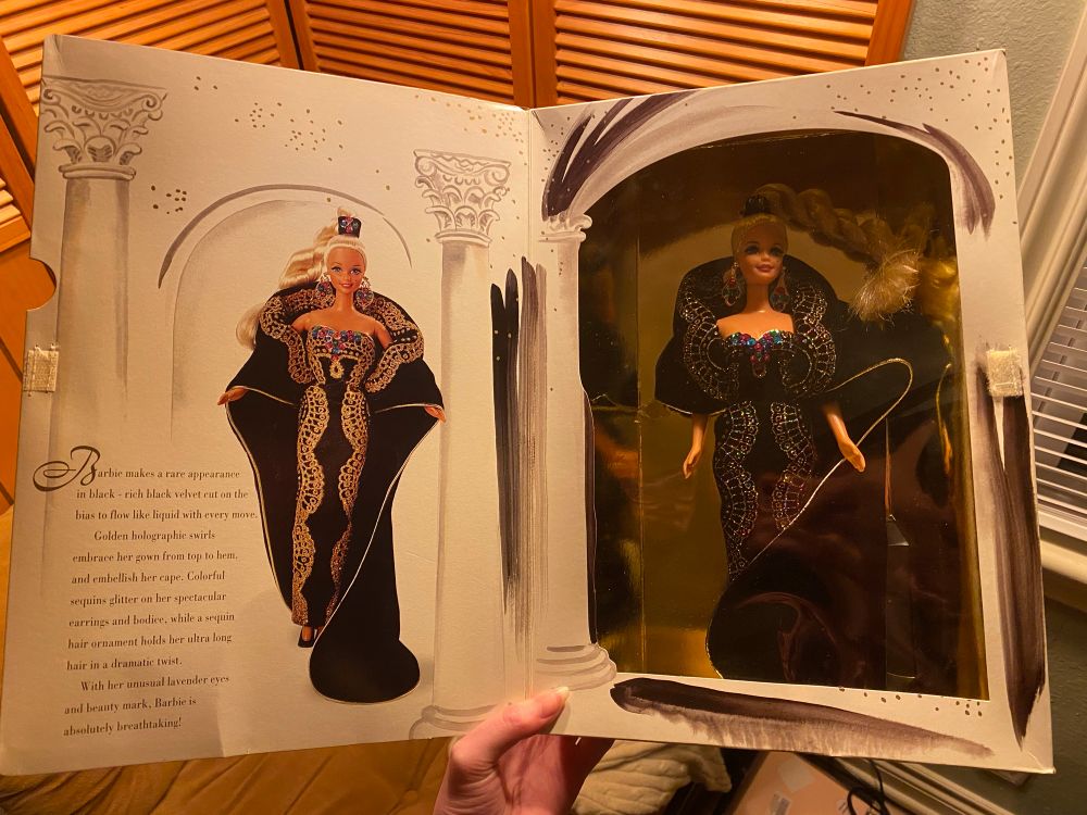Inside of the midnight gal Barbie box cover with— there’s a photo of her + a paragraph of text with her backstory (I’m sorry I’m in bed I can’t read it rn without my glasses) on the bottom left. On the right the Barbie is on display in the Box through the clear plastic layer. She is gorgeous !! Wearing a black dress with jewels and jeweled embroidery and a high pony blonde braid.