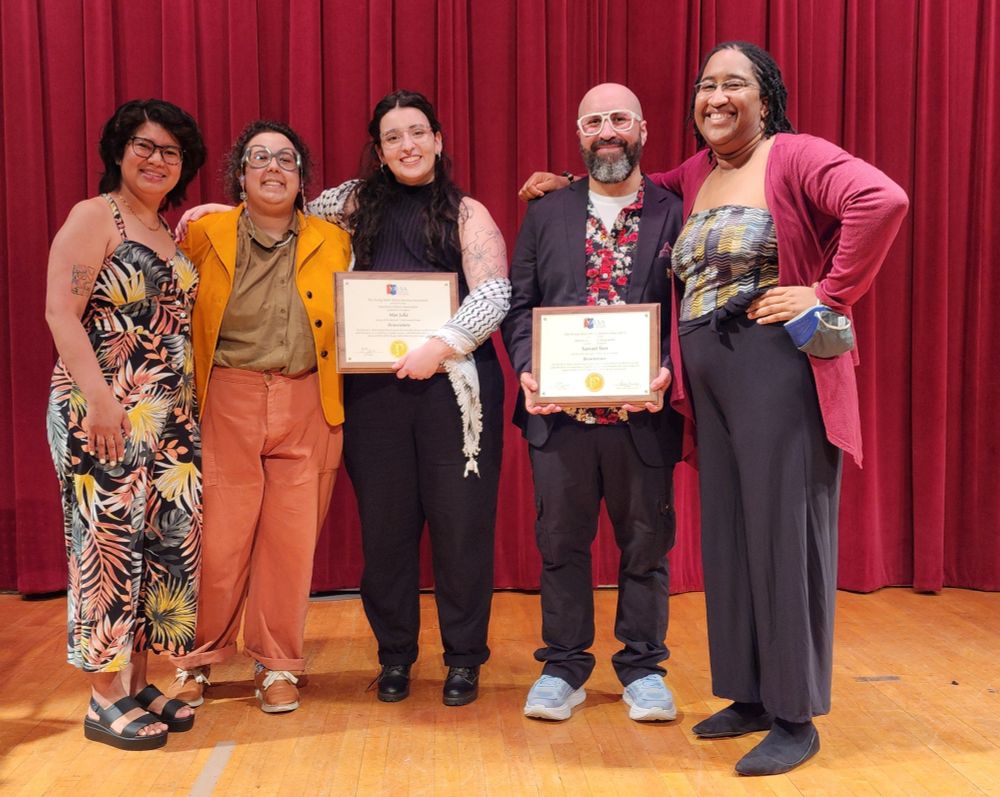 A photo of five adults of various genders with various skin tones -- from left to right, they are Mary Claire Cruz, art director; Ashanti Fortson, color designer; Mar Julia, illustrator; Samuel Teer, author; and Weslie Turner, senior editor. They stand together on a stage in front of a red curtain, most with their arms around each other, smiling at the camera. The two in the middle, Mar Julia and Samuel Teer, each hold in one hand in front of them, just below their chest, a wooden plaque with a wooden backplate and a white plate featuring the Young Adult Library Services Association (YALSA) logo above and Michael L. Printz Award seal below centered text.