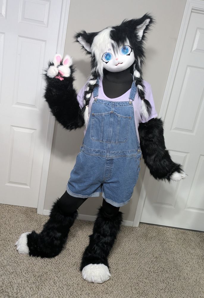 Ocean, wearing her furry Kigurumi Cookies’n’Cream, is wearing a different outfit, denim shortalls with a purple shirt! She is standing with her right paw making a peace sign.