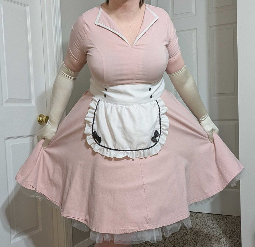 Ocean is wearing a pink 50s dress, complete with a white apron and gloves. She is performing a little curtsy, slightly lifting her dress.