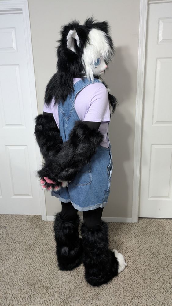 Ocean, wearing her furry Kigurumi Cookies’n’Cream, is turned with her back to the camera. She is looking over her shoulder with her paws crossed behind her back. She is wearing a different outfit, denim shortalls with a purple shirt!
