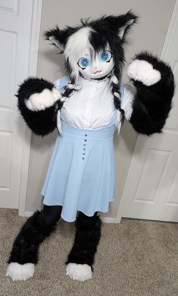 Ocean, wearing her Furry Kigurumi Cookies’n’Cream, is standing upright. Her arms are bent upwards and her paws are shifted forward. She has one leg slightly bent, with her lower paws turned slightly inward. She is wearing her usual blue skirt and white shirt.