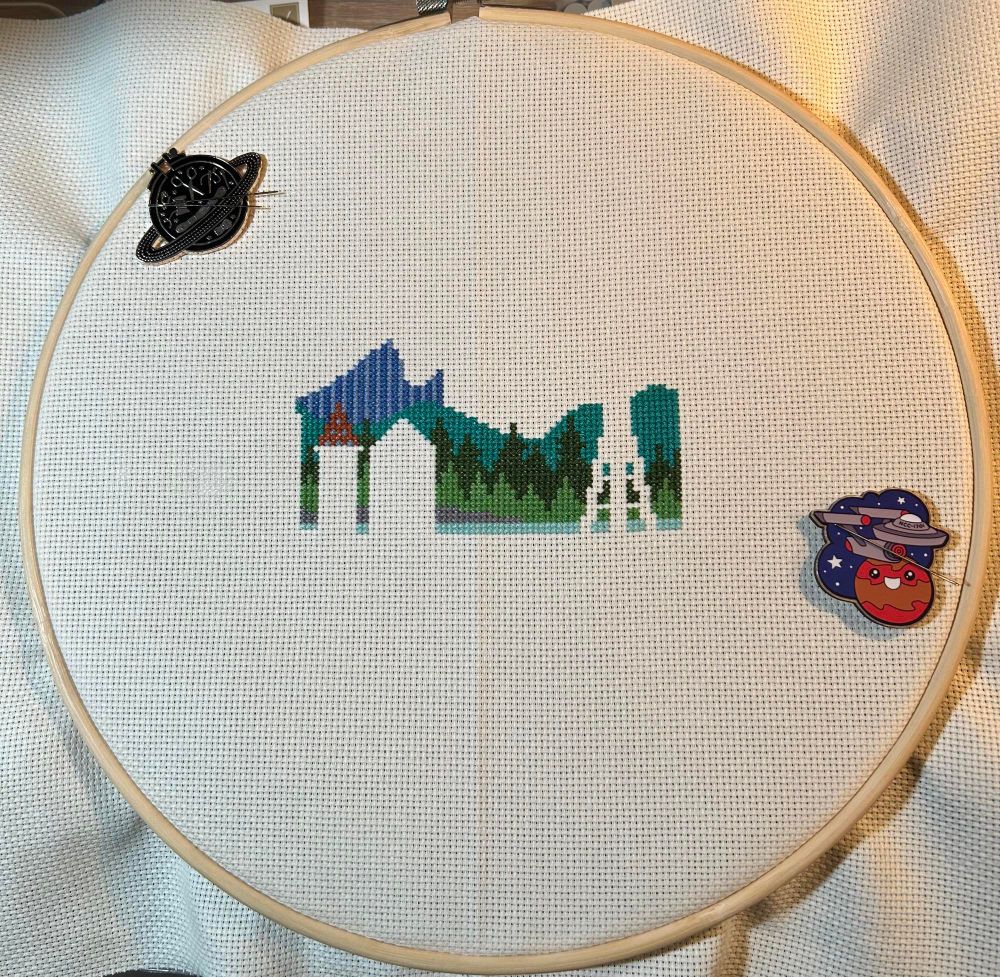 Incomplete cross stitch showing a landscape with forest and hills and the blank silhouette of buildings