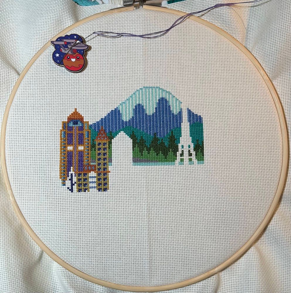 Incomplete cross stitch of a whimsical Seattle skyline.  Mountain in the background, buildings being stitched in the foreground.