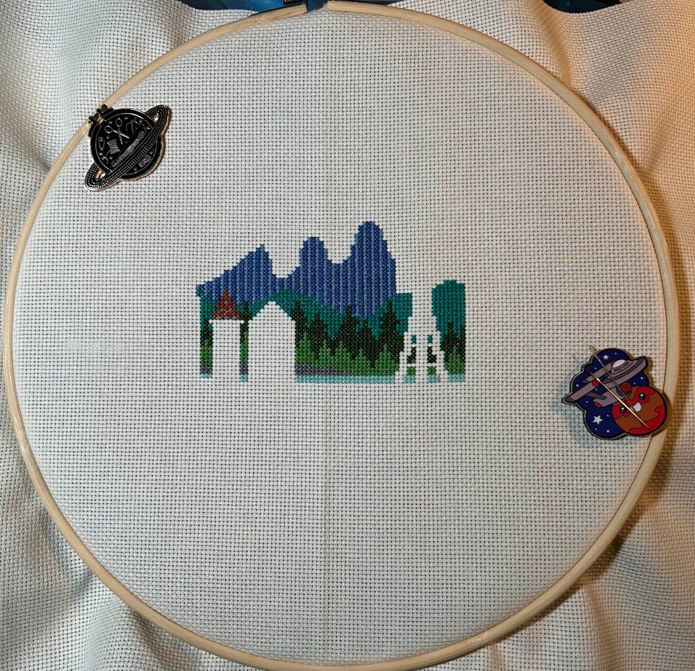 Incomplete cross stitch of a Pacific Northwest stylized landscape with blank silhouette of Seattle skyline outlined in foreground.