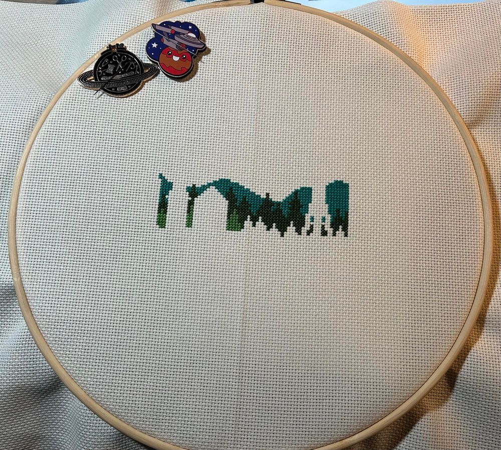 Incomplete cross stitch showing foreground tree line with hills in the distance