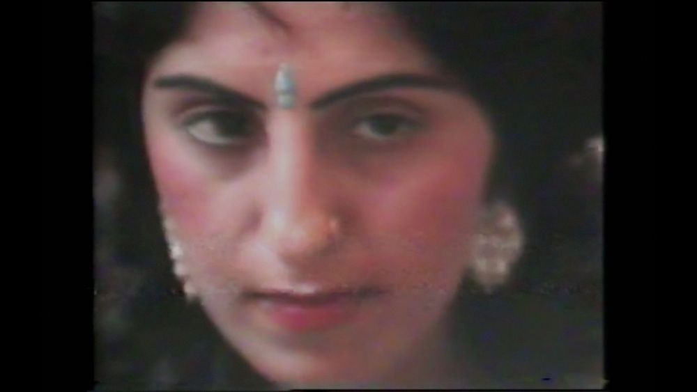 An image from Warm Shadows. It is a close up image of a person with dark hair and eyes wearing a bindi and large gold earrings. The image has a grainy vintage look. 