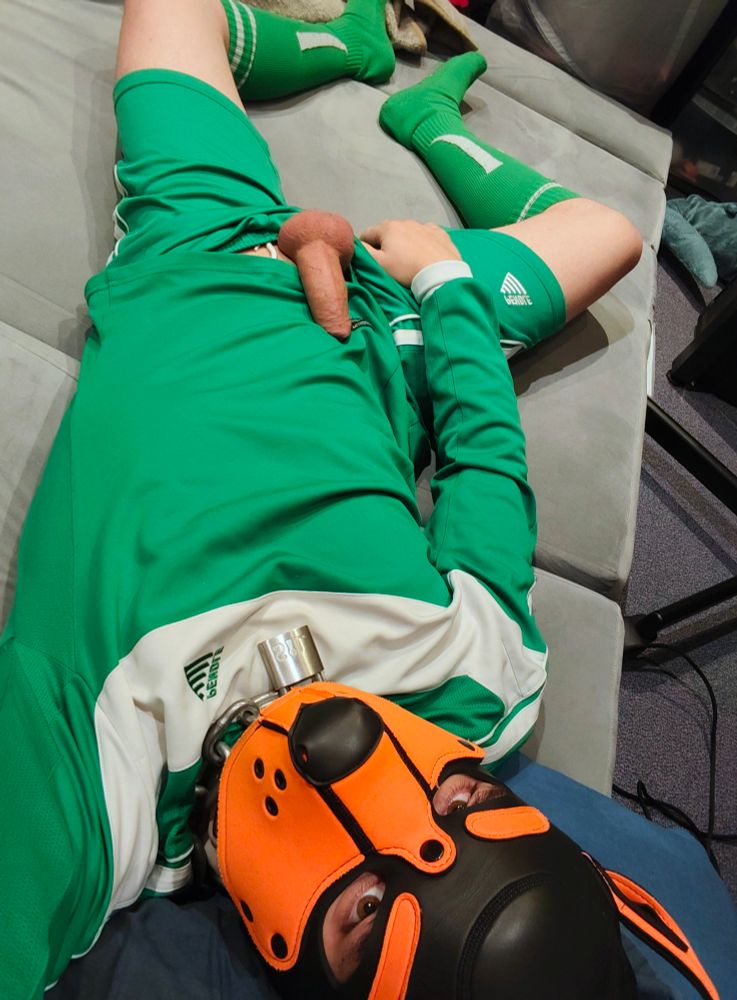 A person (it/its) lying in bed, wearing an orange pup hood and green football kit. Its hard cock is fully out over its jersey.