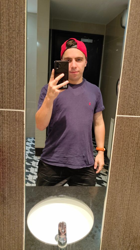 A person (it/its) taking a selfie of themself in a mirror above a sink in a bar bathroom. Despite it being 5ft3in, the sink being as low as it is makes it look larger than usual.