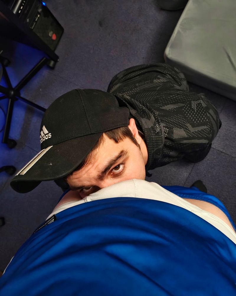 A person (it/its; @knot.ceo) with its face pressed into someone's (he/him; @astra.blue) crotch who has he shorts down slightly. @knot.ceo is wearing trackies and a black cap facing forwards; @astra.blue is wearing a blue football kit and white briefs.