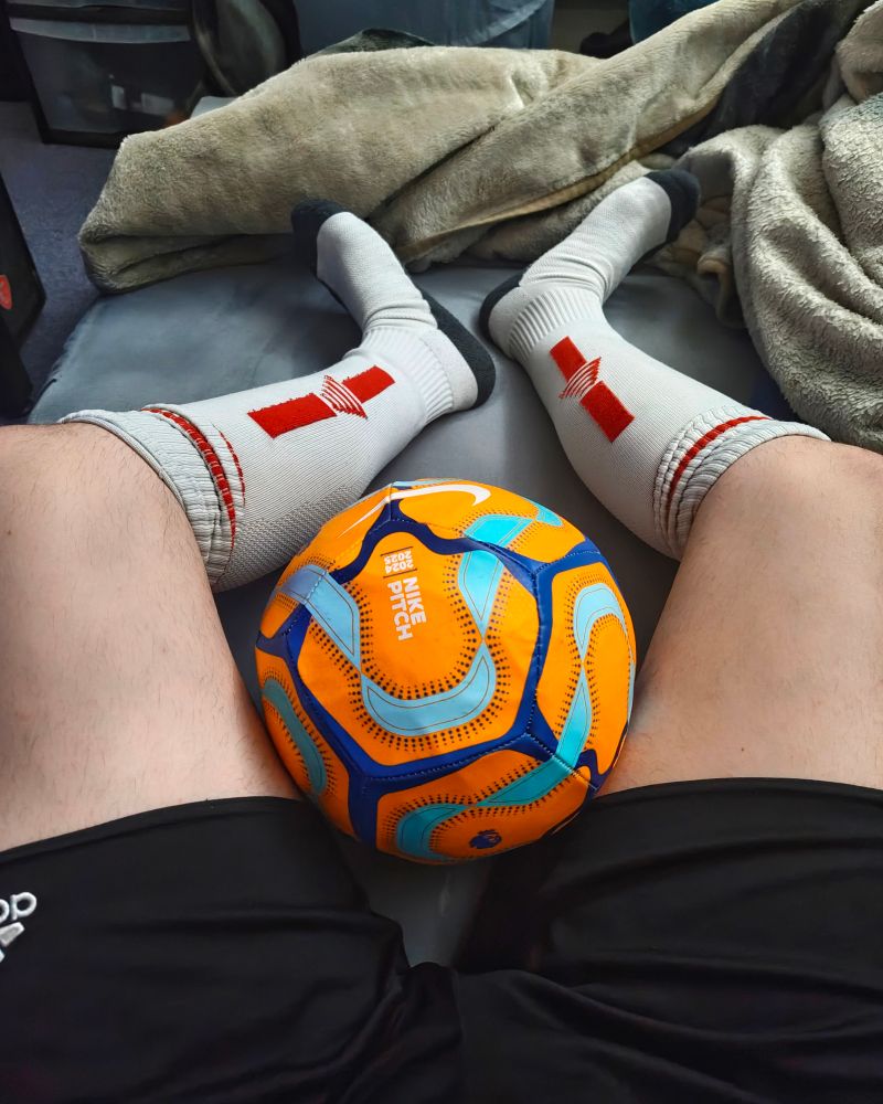 A person (it/its) lying on a bed, wearing white/red football socks, with an orange football between its crotch.