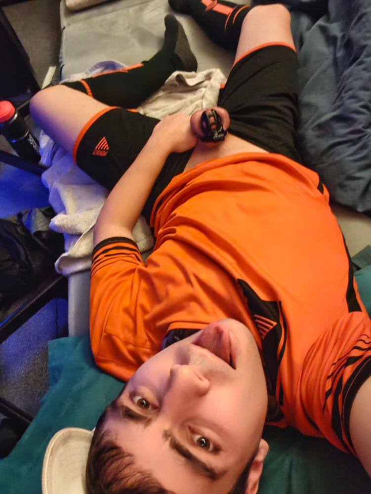 A person (it/its) lying on a bed with its shorts down, holding under the base of its cock which is locked in chastity. It's wearing an orange/black football kit, and white cap facing forwards.