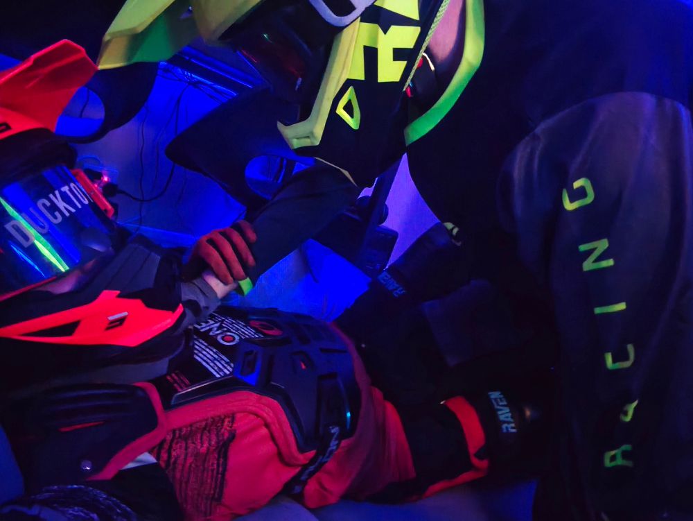 Two people embracing each other on a bed in full Motocross gear, staring straight into each other's tinted goggles.