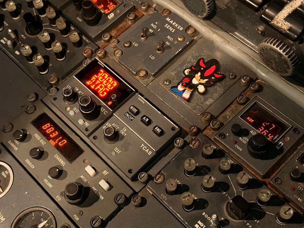 The centre pedestal of a dash 8 showing audio panels, a HF radio, a transponder, TCAS, marker switches, and a shadow the hedgehog vinyl bag tag. 