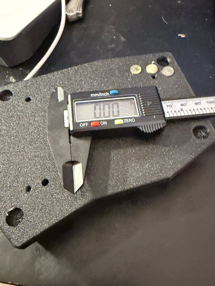 A small black digital caliper with an LCD screen reading “0.00” rests on a textured black pla plate with multiple drilled holes of varying sizes. The caliper’s jaws are closed, and its silver measuring scale extends to the right. The background shows a cluttered dark work surface with light scratches and a white cable looping from the top edge.