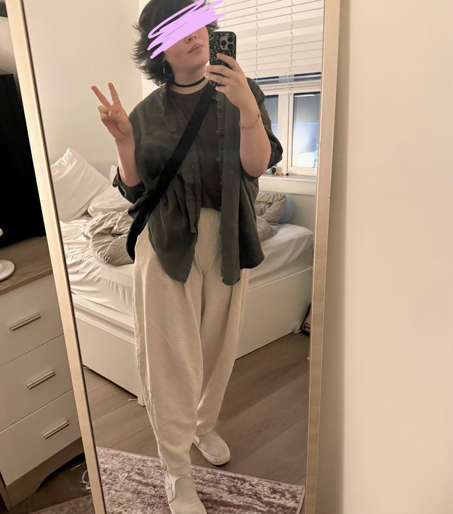 sara standing in her bedroom in front of a standing mirror, taking a photo on her front camera. she’s wearing light tan baggy pants and a baggy open green top over a brown cropped t-shirt (no midriff is showing though! you fucking pervert). she has light slip-on vans that more or less match the rest of the fit.  she’s also wearing a black cross-body bag from baggu.