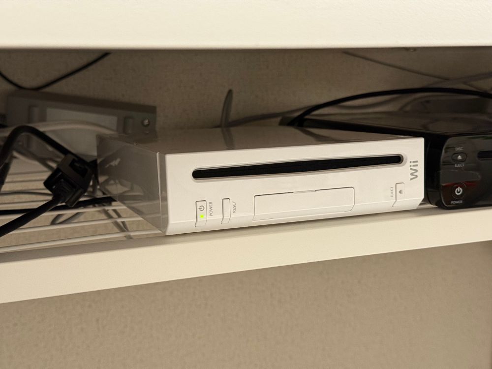 a nintendo wii on a messy shelf with an ethernet to USB adapter plugged into it, with a wii u next to it being mostly out of frame
